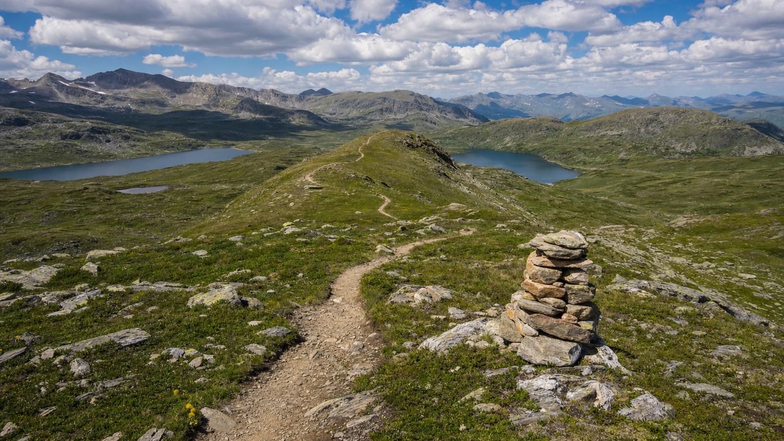 Walkers on a stony mountain path above a lake, with high peaks and late snow patches in the background